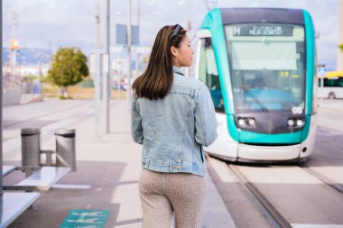 Tramvay durağında duran kot ceketli bir kadın yaklaşan bir tramvaya bakıyor. Modern bir şehir ortamında toplu taşıma için bekliyor. Yolculuk ya da yolculuk için hazır..