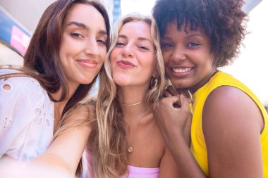 Çeşitli özelliklere sahip üç genç kadından oluşan bir grup, şehir ortamında selfie çekerken gülümsüyor. Sıradan yaz kıyafetleri giyip şehirde eğlenceli bir anın tadını çıkarıyorlar..