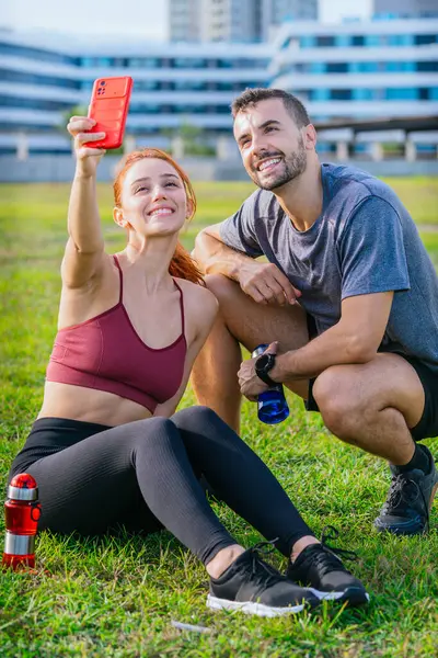 Gülümseyen genç adam ve kadın çimenlerde oturup egzersiz yaptıktan sonra selfie çekiyorlar. Spor kıyafetleri giyiyorlar ve ellerinde su şişeleri var. Şehir parkında rahat bir anın tadını çıkarıyorlar..