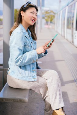 Kot ceketli şık genç bir kadın tramvay durağında oturuyor, akıllı telefonunu tutuyor. O, rahat bir ifadeyle yan tarafa bakar. Modern şehir ulaşımı ve şehir hayatı kavramı.