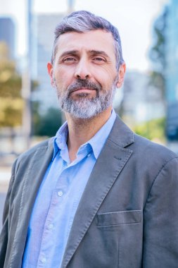 Açık havada saçı rüzgarda uçuşan gülümseyen olgun bir adamın yakın çekimi. Mutluluk ve güven saçıyor, bulanık bir şehir geçmişiyle çevrili..