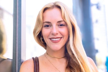 Sarışın bir kadın kollarını kavuşturmuş, şık bir yaz kıyafeti giyiyor. Bir şehir ortamında gündelik ve modaya uygun bir şekilde çekilmiş, modern zarafet sergileniyor..