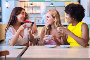 Üç farklı kadından oluşan bir grup bir masada oturuyor, ellerinde kahve fincanları, ve sıcak bir kafede birlikte gülüyorlar. Sıcak ve neşeli atmosfer, dostluğu ve bağlantıyı simgeliyor..