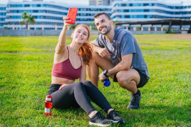 Gülümseyen genç adam ve kadın çimenlerde oturup egzersiz yaptıktan sonra selfie çekiyorlar. Spor kıyafetleri giyiyorlar ve ellerinde su şişeleri var. Şehir parkında rahat bir anın tadını çıkarıyorlar..
