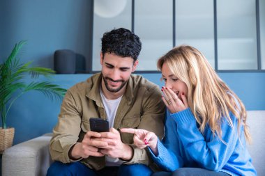 Şaşırmış ve mutlu bir çift bir kanepede oturuyor, akıllı telefona bakıp gülüyorlar. Parlak ve modern bir ev, paylaşılan keyifli anın tadını çıkarıyor. 