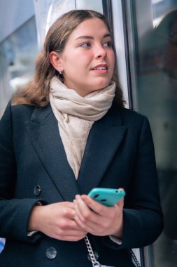 Siyah ceketli ve atkılı düşünceli genç bir kadın trenin camından dışarı bakarken elinde akıllı bir telefon tutuyor. Yansıması derinlik, yolculuk, bekleme ve tefekkür katıyor.. 