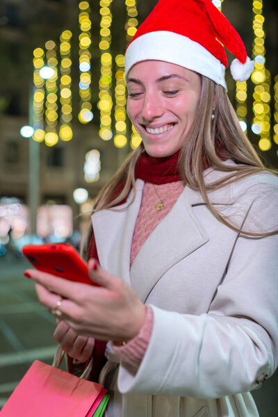 Женщина в шляпе Санты улыбается, глядя на свой телефон на праздничной городской улице ночью, в окружении светящихся праздничных огней. Перфект к Рождеству, технологии и новые темы.