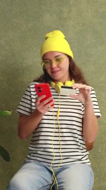 Sarı bereli, güneş gözlüklü, elinde akıllı telefon ve kredi kartıyla gülümseyen son moda genç bir kadın. Sahne çevrimiçi alışveriş ve dijital işlemleri vurguluyor. 