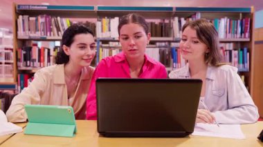 Kütüphanedeki üç genç kadın, şaşırmış ifadeleriyle bir dizüstü bilgisayarın etrafında toplandılar. Sahne, kitap raflarıyla çevrili akademik bir ortamda takım çalışması, merak ve öğrenmeyi ön plana çıkarıyor..
