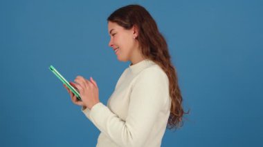 Beyaz kazaklı, yeşil tabletle mavi arka planda etkileşime giren mutlu genç bir kadın. Modern teknolojiyi kullanarak keyif ve odaklanmayı ifade etmek. 