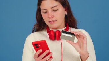 Kırmızı kulaklık takan, elinde bir kredi kartı ve akıllı telefon tutan genç bir kadın online alışveriş yaparken odaklanmış görünüyor. Mavi arka plan dijital ödeme kavramını vurguluyor. 