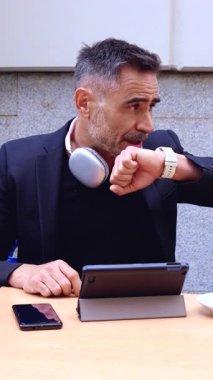 Siyah takım elbiseli profesyonel bir işadamı kol saatine bakarken akıllı telefonuyla konuşur. İfadesi, yoğun bir çalışma ortamında acil durum ve zaman yönetimi öneriyor.. 