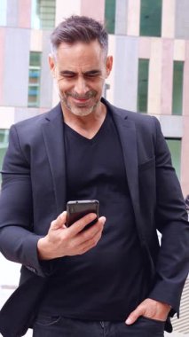 Siyah takım elbiseli kendine güvenen bir işadamı akıllı telefonunu şehir ortamında dostça bir gülümsemeyle uzatır. Onun jesti teknoloji, iş ve dijital iletişimi iletir.. 