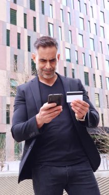 Siyah takım elbiseli profesyonel bir işadamı şehir ortamında akıllı telefonunu kullanırken bir kredi kartı tutuyor. Odaklanmış ifadesi online bankacılık ya da dijital ödeme öneriyor. 