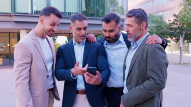 İş dünyasından dört iş adamı resmi ve günlük giysiler içinde dışarıda toplandı, akıllı telefona dikkatle baktılar, kentsel bir ortamda işle ilgili bilgileri tartıştılar.
