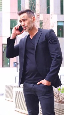 Siyah takım elbiseli profesyonel bir işadamı şehir ortamında dururken akıllı telefonuyla konuşur. Onun ciddi ifadesi önemli bir iş görüşmesi öneriyor.. 