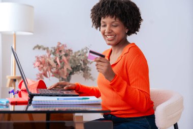 Afrikalı Amerikalı öğrenci evde dizüstü bilgisayar kullanırken kredi kartıyla çevrimiçi ödeme yapıyor. Kitaplı, kırtasiyeli ve şık dekorlu modern çalışma alanı. E-ticaret, dijital işlem ve finansman