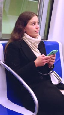 Siyah ceketli ve bej eşarplı neşeli genç bir kadın modern bir metro treninde oturmuş akıllı telefonuna bakıp gülüyor. Seyahat sırasında eğlenceli bir anın tadını çıkarıyor.. 
