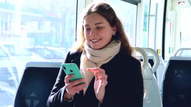 Siyah ceketli ve bej eşarplı neşeli genç bir kadın modern bir trenin içinde oturmuş akıllı telefonuna bakıp gülüyor. Seyahat sırasında eğlenceli bir anın tadını çıkarıyor.. 