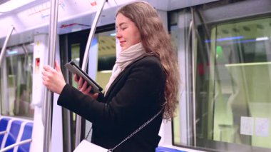  Modern bir metro treninde dururken siyah paltolu ve bej eşarplı şık bir kadın bir e-okuyucuda okuyor. Yolda giderken bir direğe tutunarak dengede duruyor..