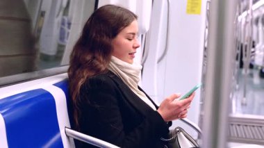 Siyah ceketli ve bej eşarplı neşeli genç bir kadın modern bir metro treninde oturmuş akıllı telefonuna bakıp gülüyor. Seyahat sırasında eğlenceli bir anın tadını çıkarıyor.. 
