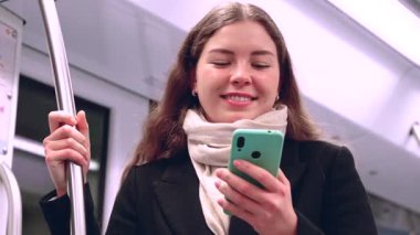 Kafkasyalı genç bir kadın akıllı bir telefon tutuyor ve metroda duruyor, sırığı tutarken hafifçe gülümsüyor, toplu taşıma sırasında siyah bir ceket ve eşarp takıyor..