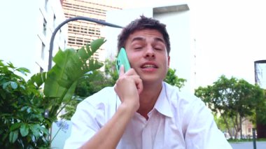 Açık bir kafede otururken telefonla konuşan beyaz gömlekli bir genç adam. Dijital iletişimle rahat ve modern bir yaşam tarzı.. 