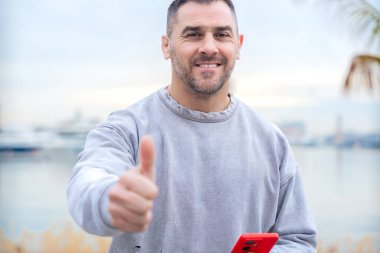 Elinde kırmızı bir akıllı telefon tutan gülümseyen beyaz adam, limanda palmiye ağaçlarıyla baş parmaklarını gösteriyor, gri bir kazak giyiyor, neşeli ve dostça bir ifadeyle onay ve pozitifliğini dile getiriyor..