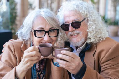 Gri saçlı ve gözlüklü yaşlı bir çift kahvenin tadını çıkarıyor. Kahverengi paltolarını giyip, fincanlarına kadeh kaldırarak gülümsüyor, zarif şehir ortamında neşeli bir anı paylaşıyorlar. Sıcak ve misafirperver bir atmosfer.