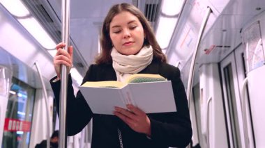 Modern bir metro treninde duran siyah paltolu ve bej eşarplı odaklanmış genç bir kadın metal bir direğe tutunarak kitap okuyor. Seyahat sırasında sakin bir anın tadını çıkarıyor..