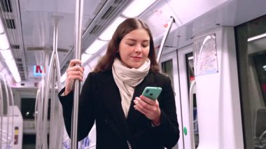 Kafkasyalı genç bir kadın akıllı bir telefon tutuyor ve metroda duruyor, sırığı tutarken hafifçe gülümsüyor, toplu taşıma sırasında siyah bir ceket ve eşarp takıyor..