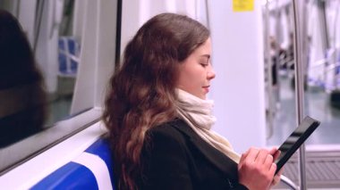 Modern bir metro treninde oturan siyah paltolu ve bej eşarplı odaklanmış genç bir kadın bir e-okuyucuda okuyor. Her gün işe gidip gelirken sakin bir anın tadını çıkarıyor.. 