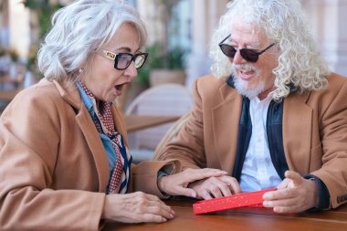 Dışarıda bir kafede partnerinden hediye alan son sınıf öğrencisi heyecanlı bir kadın. Yaşlı çift, bu görüntüyü aşk, sürpriz ve ilişki temaları için mükemmel kılan neşeli ve sevgi dolu bir anı paylaşıyor.