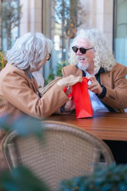 Dışarıda bir kafede partnerinden hediye alan son sınıf öğrencisi heyecanlı bir kadın. Yaşlı çift, bu görüntüyü aşk, sürpriz ve ilişki temaları için mükemmel kılan neşeli ve sevgi dolu bir anı paylaşıyor.