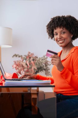 Afrikalı Amerikalı bir öğrenci evde telefon kullanırken kredi kartıyla online ödeme yapıyor. Kitaplı, kırtasiyeli ve şık dekorlu modern çalışma alanı. E-ticaret, dijital işlem ve finans