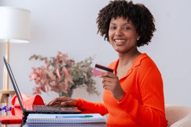 Afrikalı Amerikalı bir öğrenci evde telefon kullanırken kredi kartıyla online ödeme yapıyor. Kitaplı, kırtasiyeli ve şık dekorlu modern çalışma alanı. E-ticaret, dijital işlem ve finans
