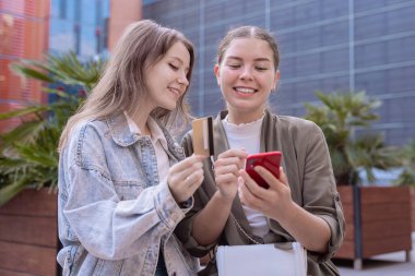 Açık havada alışveriş yapmak için akıllı telefon ve kredi kartı kullanan gülümseyen iki beyaz kadın. Modern yaşam tarzı, dijital ödeme ve teknoloji konsepti kentsel ortamda doğal gün ışığı ile.