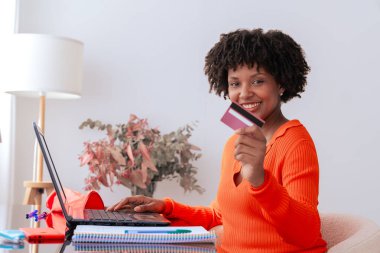 Afrikalı Amerikalı bir öğrenci evde telefon kullanırken kredi kartıyla online ödeme yapıyor. Kitaplı, kırtasiyeli ve şık dekorlu modern çalışma alanı. E-ticaret, dijital işlem ve finans