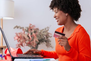 Afrikalı Amerikalı öğrenci evde dizüstü bilgisayar kullanırken kredi kartıyla çevrimiçi ödeme yapıyor. Kitaplı, kırtasiyeli ve şık dekorlu modern çalışma alanı. E-ticaret, dijital işlem ve finansman