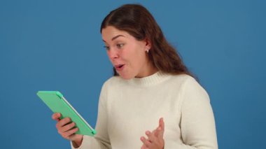 Yeşil tablet kullanırken beyaz kazaklı neşeli genç beyaz kadın başarıyı kutluyor. Heyecanlı bir ifade, neşeli bir tepki ve dijital ortamda mavi bir arkaplan karşısında modern bir yaşam tarzı.