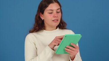 Beyaz kazaklı genç beyaz kadın yeşil tablet kullanıyor, ekrana odaklanıyor. Düşünceli ifade, modern teknoloji kullanımı ve mavi arka plana karşı dijital etkileşim.