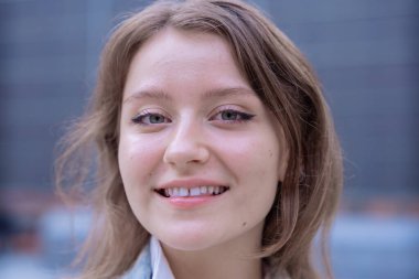Genç beyaz iş kadını kameraya ışıltılı bir gülümsemeyle bakıyor. Kendine güven, mutluluk ve pozitiflik yayarak, onu yaşam tarzı, moda ve kişisel markalaşma konuları için ideal bir imaj haline getiriyor.