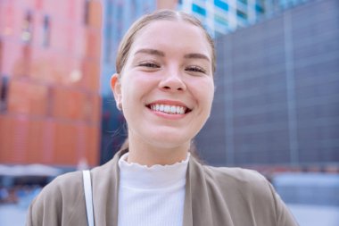 Genç beyaz iş kadını kameraya ışıltılı bir gülümsemeyle bakıyor. Kendine güven, mutluluk ve pozitiflik yayarak, onu yaşam tarzı, moda ve kişisel markalaşma konuları için ideal bir imaj haline getiriyor.