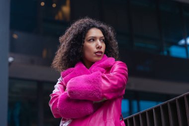 Pembe palto giyen kıvırcık saçlı zarif dansçı şehir ortamında hareketini ve enerjisini ifade ediyor. Modern moda, çağdaş dans ve sanatsal performans şehrin arka planına karşı..