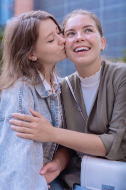 İki mutlu Kafkasyalı kadın, yanaklarından öpücükler kondurarak sevgilerini ifade ediyorlar. Arkadaşlığın, sevginin ve güvenin sembolü. Doğal ışık ve neşeli ifadelerle modern bir şehir ortamında..