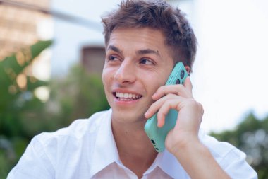 Açık hava telefon görüşmesi sırasında akıllı telefondan konuşan mutlu genç bir adam. Gülümsüyor ve başka tarafa bakıyor, modern ve rahat kentsel bir ortamda, güneş ışığıyla pozitif bir konuşma öneriyor..