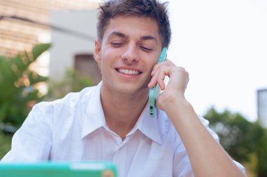 Açık hava telefon görüşmesi sırasında akıllı telefondan konuşan mutlu genç bir adam. Gülümsüyor ve başka tarafa bakıyor, modern ve rahat kentsel bir ortamda, güneş ışığıyla pozitif bir konuşma öneriyor..
