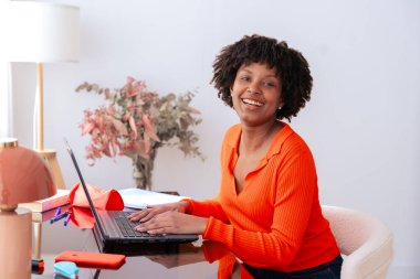 Afro-Amerikalı kadın ofis masasında oturuyor, dizüstü bilgisayarla çalışıyor ve kameraya bakıyor. Kitapları, kırtasiyesi ve dekorasyonu olan modern çalışma alanı. Uzak çalışma, üretkenlik, teknoloji ve güven.