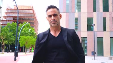 Şık, olgun, beyaz bir adam güvenli bir şekilde şehir iş alanında yürüyor. Siyah bir takım elbise giyiyor ve ciddi görünüyor. Modern arka planda yüksek binalar ve ağaçlar var.. 