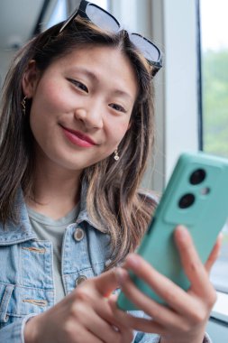 Genç Çinli bir kadın tramvayda oturuyor ve akıllı telefonuyla sosyal medyayı kontrol ederken gülümsüyor. Şehir toplu taşıma sahnesi. Yaşam tarzı, teknoloji, seyahat ve dijital bağlantı temaları için ideal.